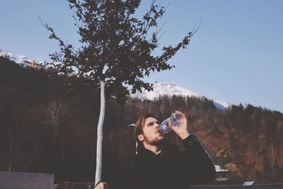 Young man drinking water against forested mountain