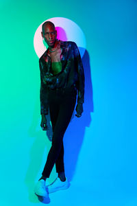 Full body of bald african american male in stylish wear standing looking at camera at wall with glowing light in studio