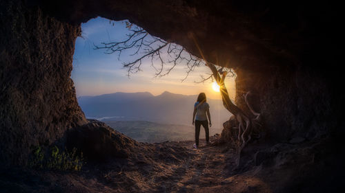 Rear view of woman standing on rock in cave against sky during sunset