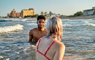 Young black male looking at girlfriend while standing in foamy sea waves on summer weekend day