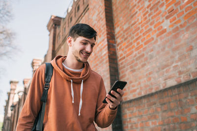 Low angle view of young man using mobile phone while standing against brick wall