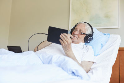 Senior man wearing headphones while using digital tablet on bed in hospital ward