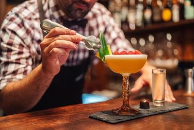 Unrecognizable barkeeper putting leaf on tropical cocktail with raspberries and pineapple near chocolate candy and liquor in pub
