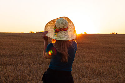 Rear view of woman wearing hat on field against sky during sunset in the wheat field countryside.