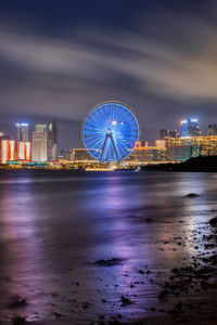 Illuminated ferris wheel by sea against sky at night