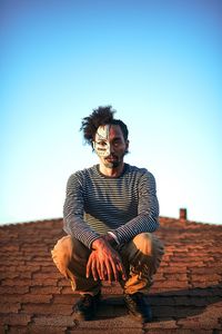 Portrait of man with painted face crouching on footpath against clear sky