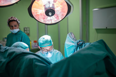 Male doctor in glasses and uniform performing surgery on patient with help of assistants in modern operating theater of hospital