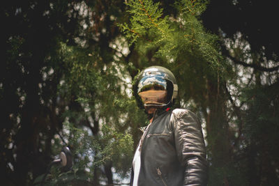 Biker wearing crash helmet and leather jacket standing against trees