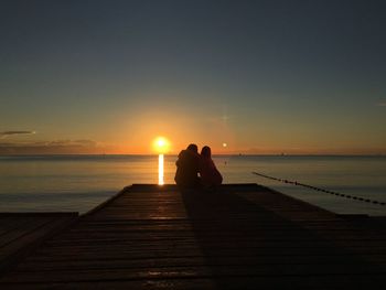 Silhouette couple sitting on pier over sea against sky during sunset