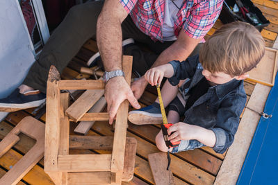 From above unshaven mature dad with attentive boy measuring wooden blocks with tape while spending time on blurred background