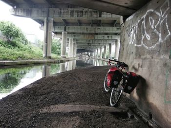 A bicycle stands against a cement wall below a bridge next to a small river