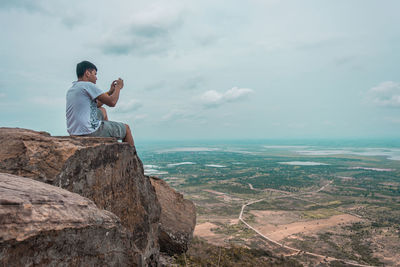 Rear view of man photographing sitting in cliff against landscape