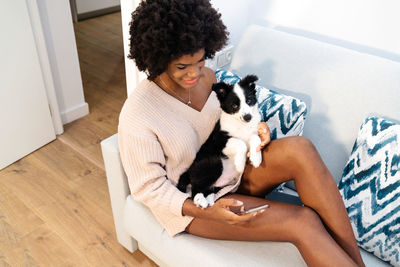 From above young black female in casual clothes sitting on sofa with smartphone on hand with cute little puppy on lap at home