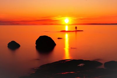 Silhouette rocks by sea against orange sky during sunset