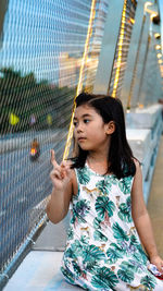 Cute girl looking away sitting by chainlink fence