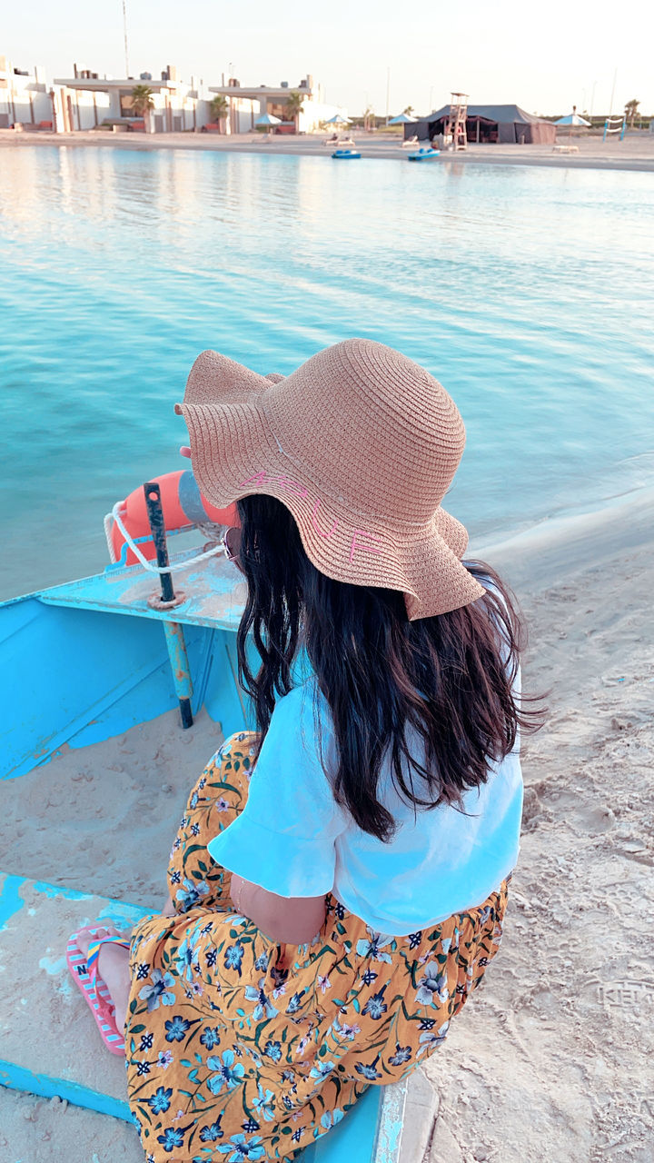 water, one person, women, sea, adult, nature, clothing, beach, hat, relaxation, land, blue, rear view, vacation, sun hat, trip, summer, fashion accessory, hairstyle, leisure activity, lifestyles, long hair, day, holiday, sitting, female, travel, full length, travel destinations, nautical vessel, outdoors, tranquility, sky, sunlight, beauty in nature, casual clothing, young adult, person, transportation, straw hat, tourism, child, brown hair