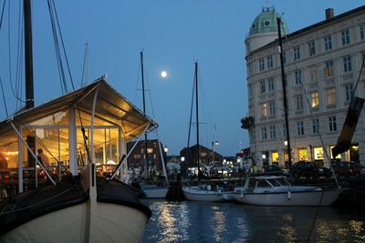 Sailboats moored on canal by illuminated city buildings against sky at dusk