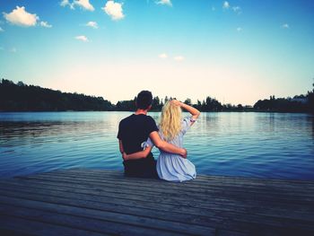 Rear view of couple sitting on pier over lake against blue sky