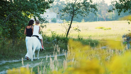 A woman and a boy are walking around the field, son is riding a pony, mother is holding a pony for