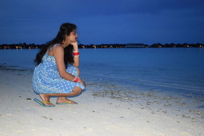 Side view of young woman looking away while crouching at beach