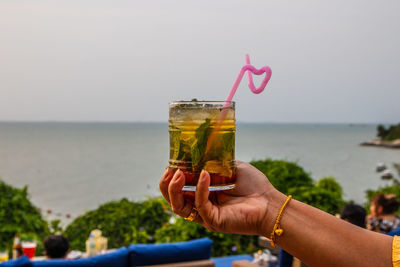 Hand holding drink at sea against sky