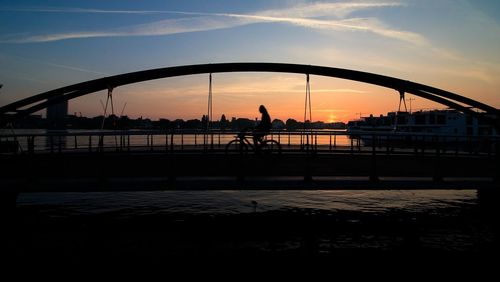 Silhouette people riding bicycle on bridge over river against sky during sunset