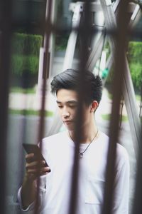 Young man using mobile phone while standing on street seen through fence