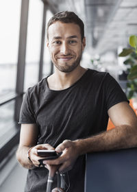 Portrait of smiling businessman holding mobile phone while leaning on cubicle in office