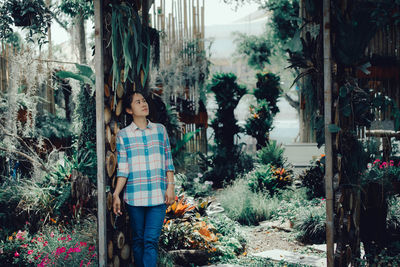 Woman looking away while standing by wall against plants in greenhouse