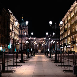Empty footpath by illuminated street lamps amidst buildings