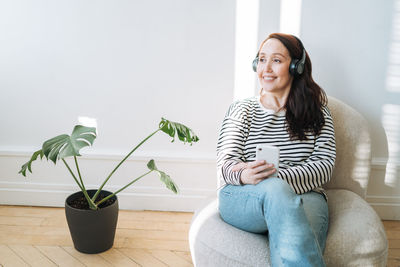 Smiling brunette woman using mobile phone, listen music in headphones in chair at the home