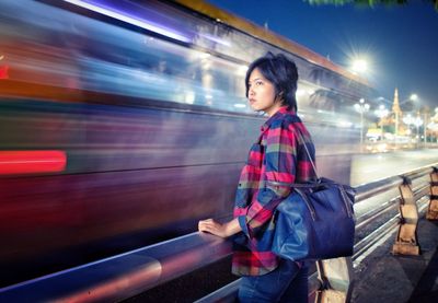 Side view of woman looking away by blurred train at night
