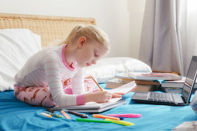 Cute girl writing in book sitting on bed at home