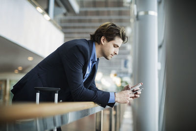 Side view of businessman using mobile phone while leaning on railing at airport