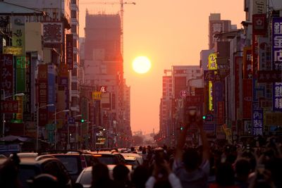 Crowd on city street by buildings against sky during sunset