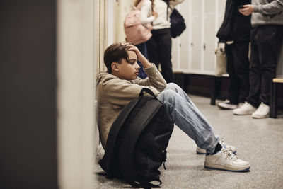 Side view of sad teenage boy sitting with backpack in school corridor