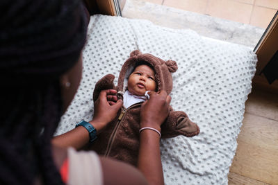 Top view of cropped unrecognizable caring african american mother with long braided hair dressing bodysuit on newborn baby lying on blanket on sofa