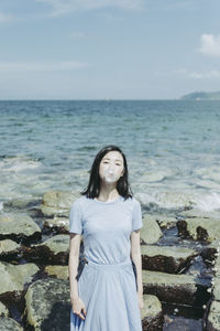Portrait of beautiful woman blowing bubble gum while standing against rock at beach