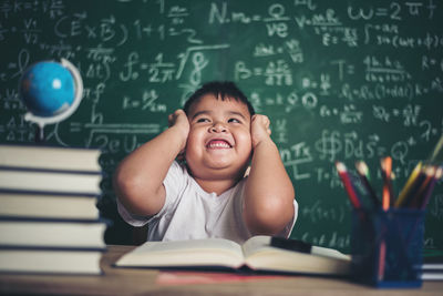 Cute boy with head in hand studying at table against blackboard