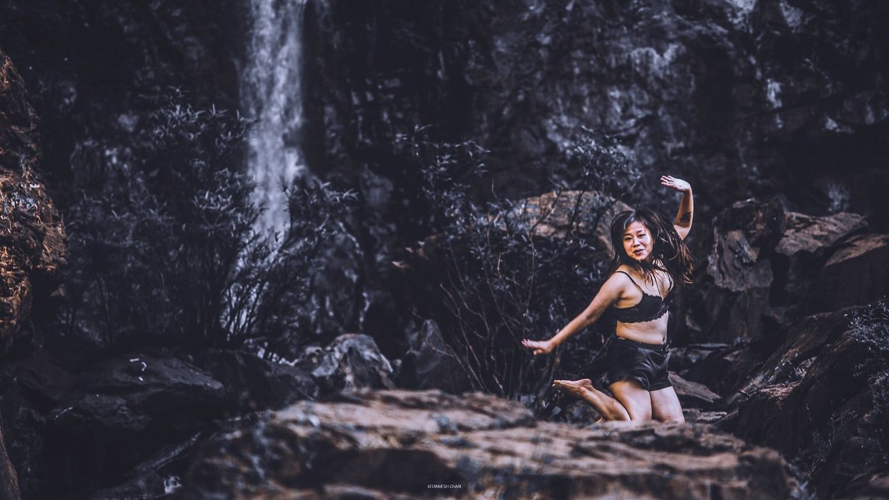 young adult, young women, rock, rock - object, nature, full length, solid, leisure activity, motion, one person, waterfall, lifestyles, forest, women, real people, water, land, sitting, adult, flowing water, beautiful woman, outdoors