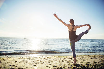 Rear view of woman practicing hand to big toe yoga pose at beach during sunny day