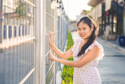 Portrait long hair asian woman in a white patterned dress standing and touching a fence at evening