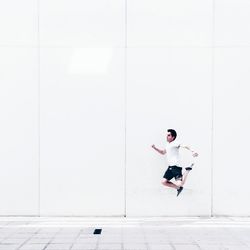 Full length of young man jumping over footpath against white wall