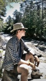 Side view of man wearing hat sitting on rock in forest during sunny day