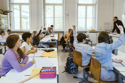 Male and female students raising hands while sitting on chairs in classroom