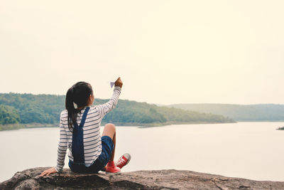 Rear view of girl playing with paper airplane while sitting on rock against lake