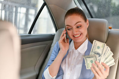 Smiling businesswoman with paper currency talking on phone while sitting in car