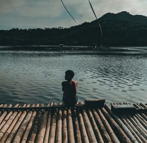 Rear view of shirtless young man crouching on pier over lake against sky during sunset