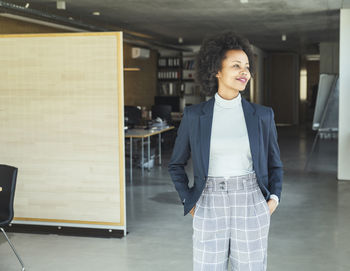 Female professional looking away while standing with hands in pockets at office