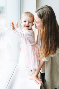 Young woman mom with long hair in cozy knitted cardigan with baby girl on hands near window at home